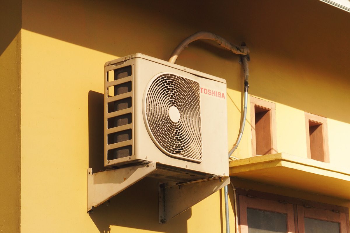 An outdoor aircon condenser on a steel wall bracket, in hard afternoon sun against a yellow building wall, its refrigerant lines running up under the eave.