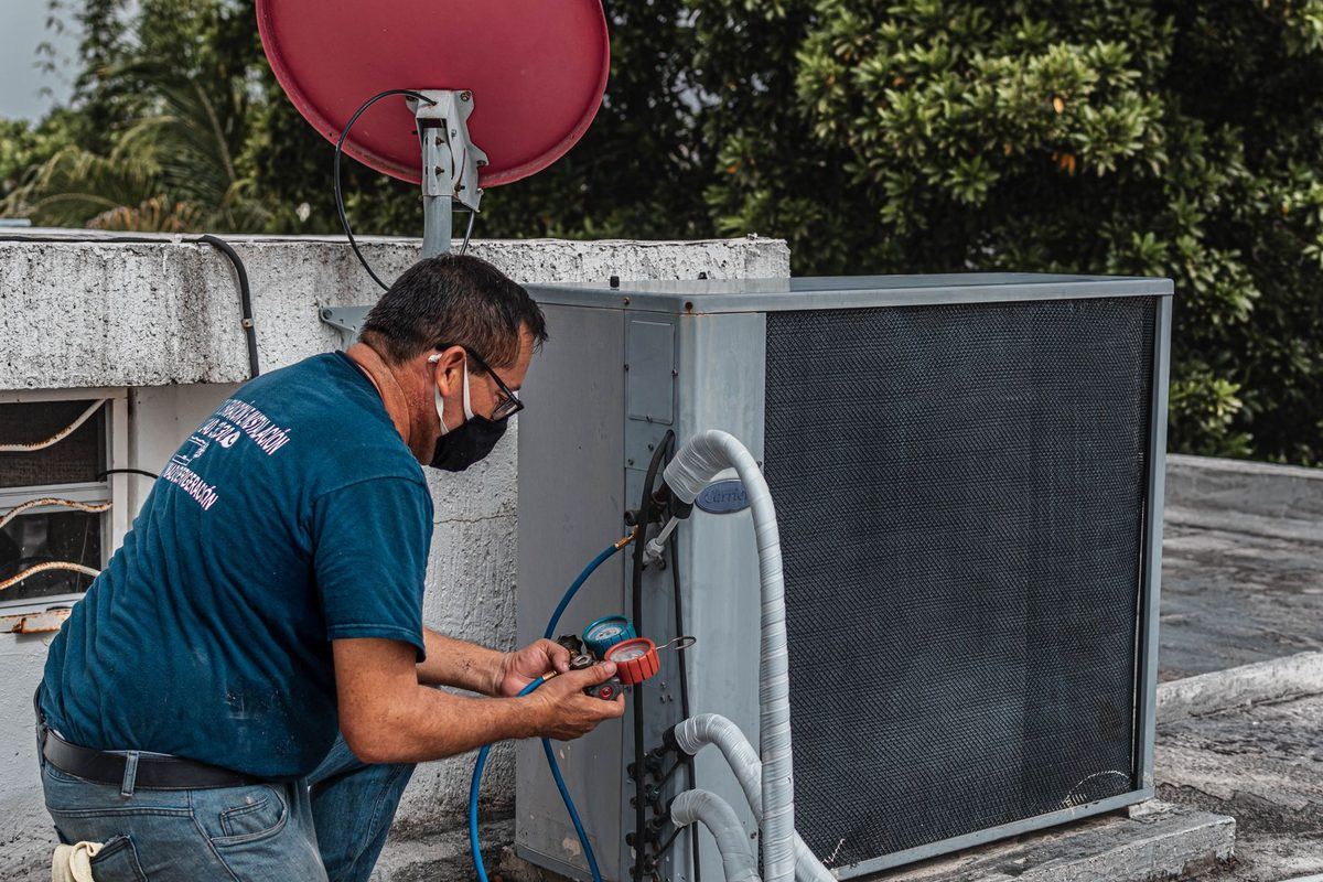 A technician in a work shirt and face mask servicing a rooftop air conditioning condenser unit with a manifold gauge, surrounded by tropical foliage.