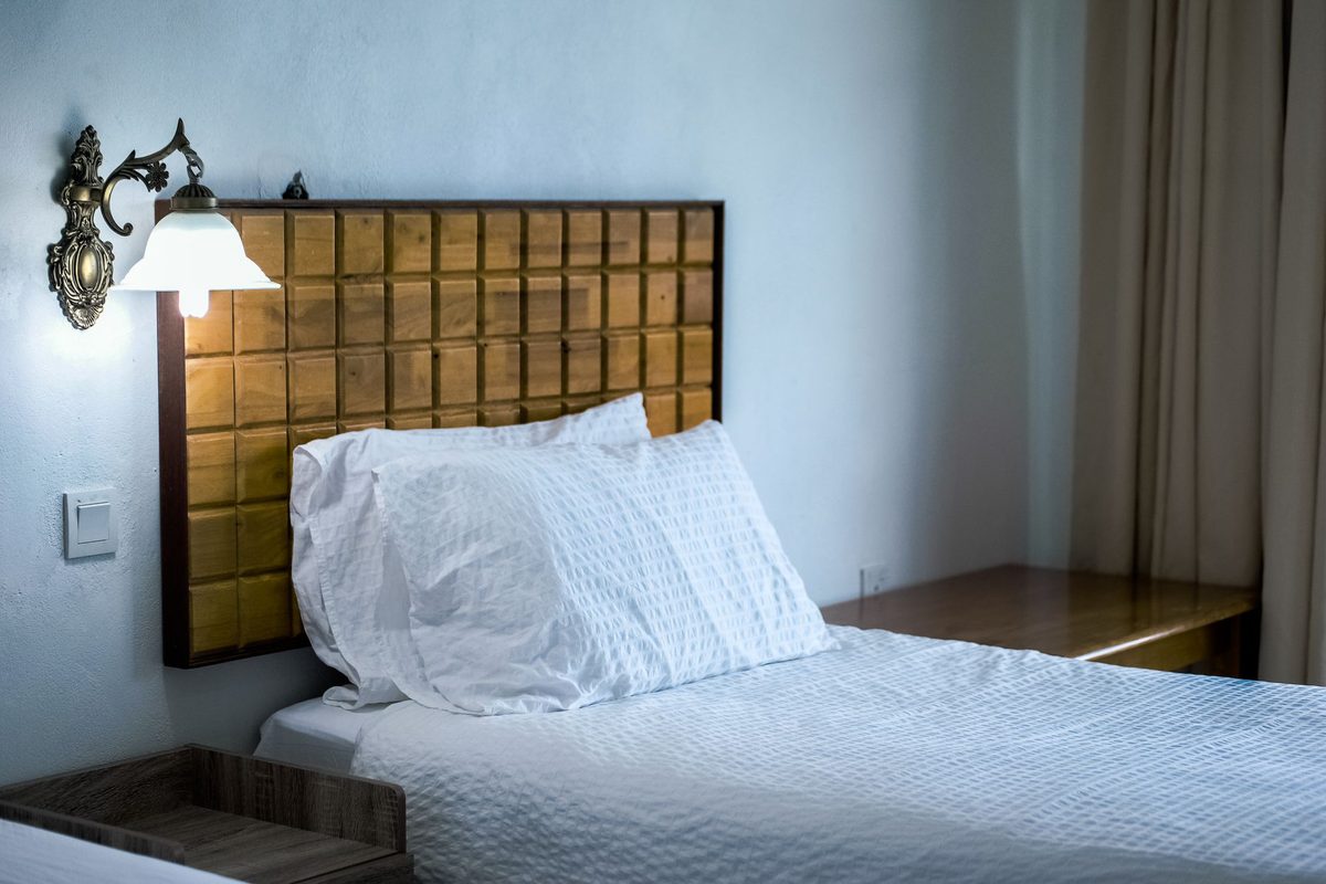 A freshly made hotel bed with white bedding beside a wooden side table, lit by a brass wall sconce above a tiled wooden headboard.