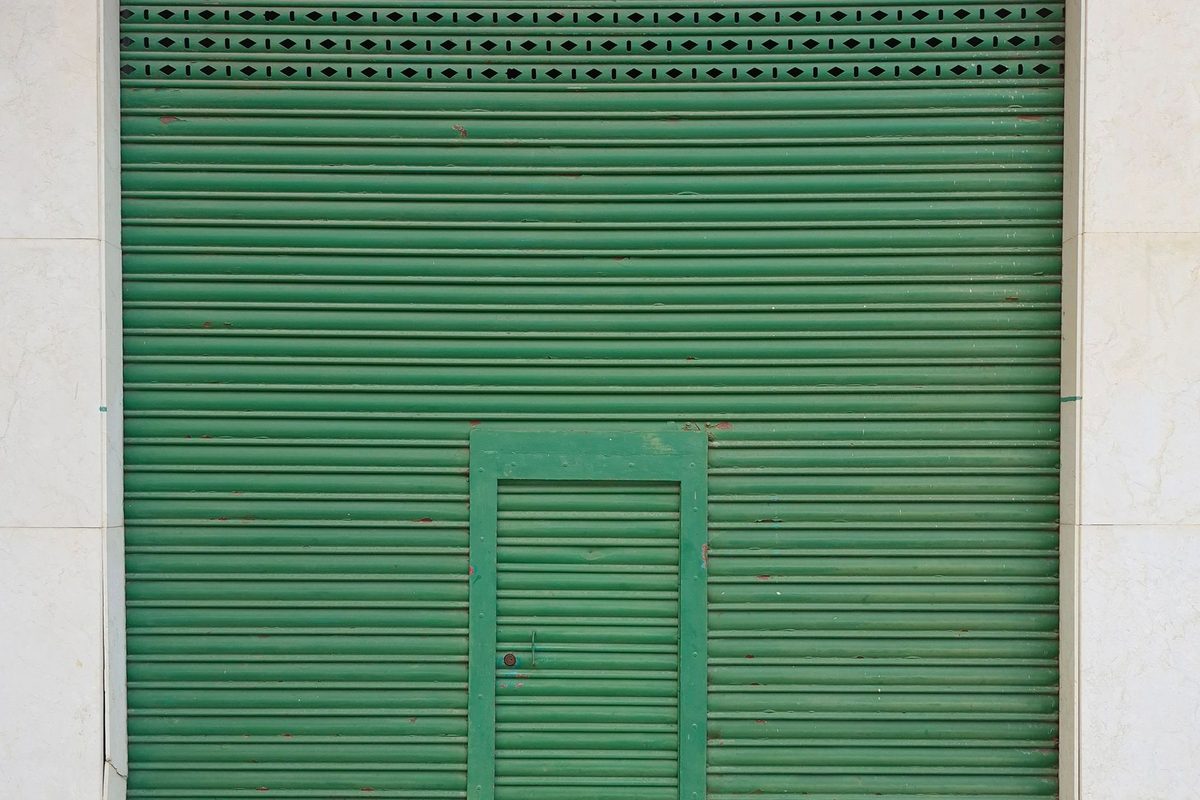 A green metal roll-down security shutter with a small inset door, pulled fully closed over a storefront between two tiled pillars.