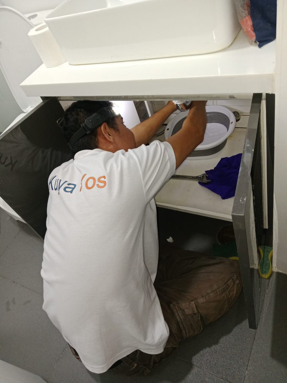 A KuyaYos plumber in uniform clearing a clogged bathroom sink drain under the vanity